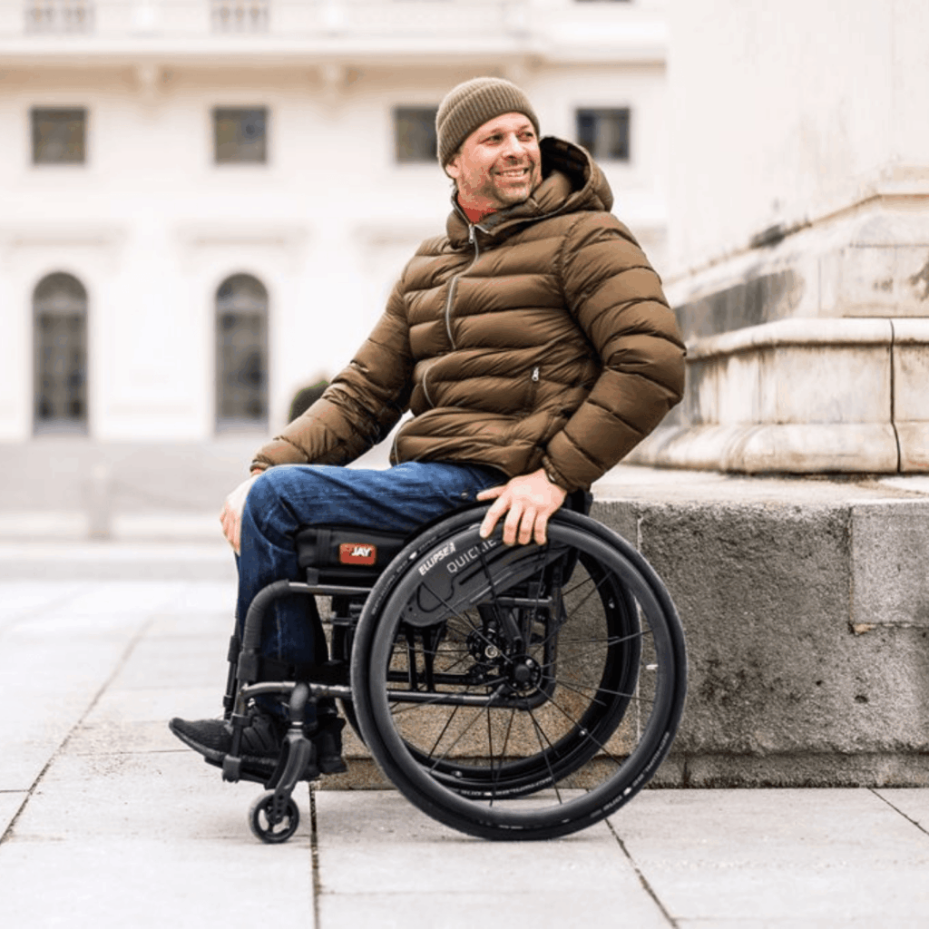 A man in a brown puffer jacket and knit beanie smiles while sitting in a black ultralight manual wheelchair. He is outdoors in an urban setting, leaning against a stone wall with classical architecture in the background.