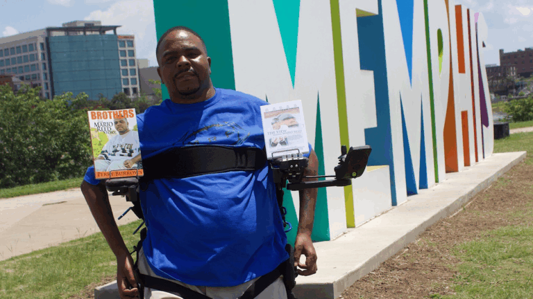 A man in a blue shirt stands in front of a large, colorful Memphis sign, wearing a harness that holds two books labeled Marios Story on a sunny day.