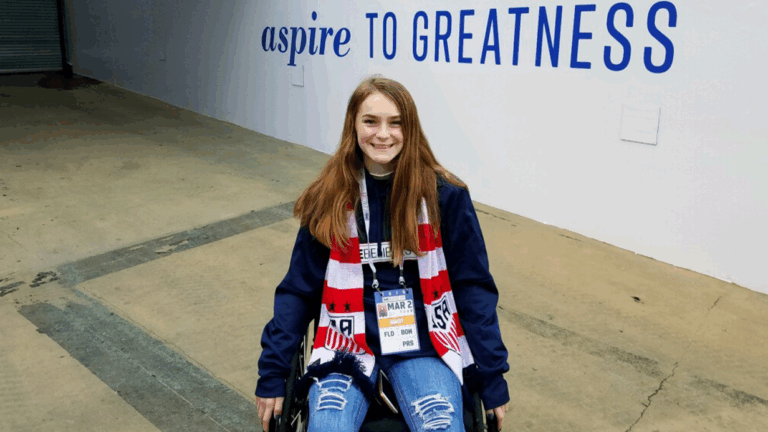 A young woman shares McKennas Story as she smiles at the camera, wearing a red, white, and blue scarf and name badges. Behind her, large blue text on the wall reads aspire TO GREATNESS.