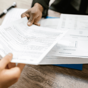 Close-up of two people exchanging paperwork across a wooden desk. The documents include printed forms and financial statements.
