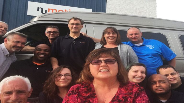 A group of thirteen smiling people, featured in Mikes Story, pose together outdoors in front of a gray van and a building with a numobi sign. The group consists of men and women of various ages.