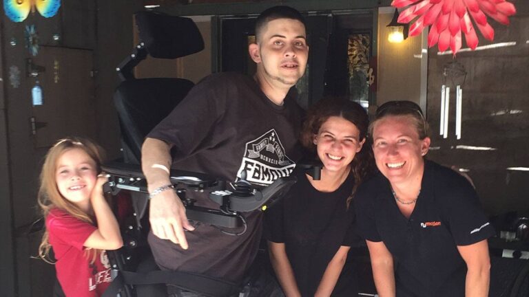 Four people smile together indoors. One man uses a wheelchair with a tray, and three women, including a young girl, stand beside him. Warm light and colorful decorations set the scene for Nicks Story in the background.