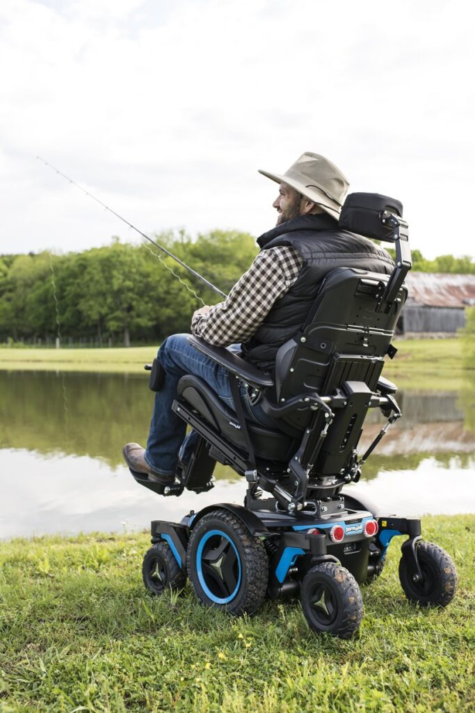 A man in a wide-brimmed hat sits in a power tilt wheelchair by a pond, fishing with a rod. He is outdoors on grass, with trees and a barn in the background, creating a peaceful and relaxed scene.