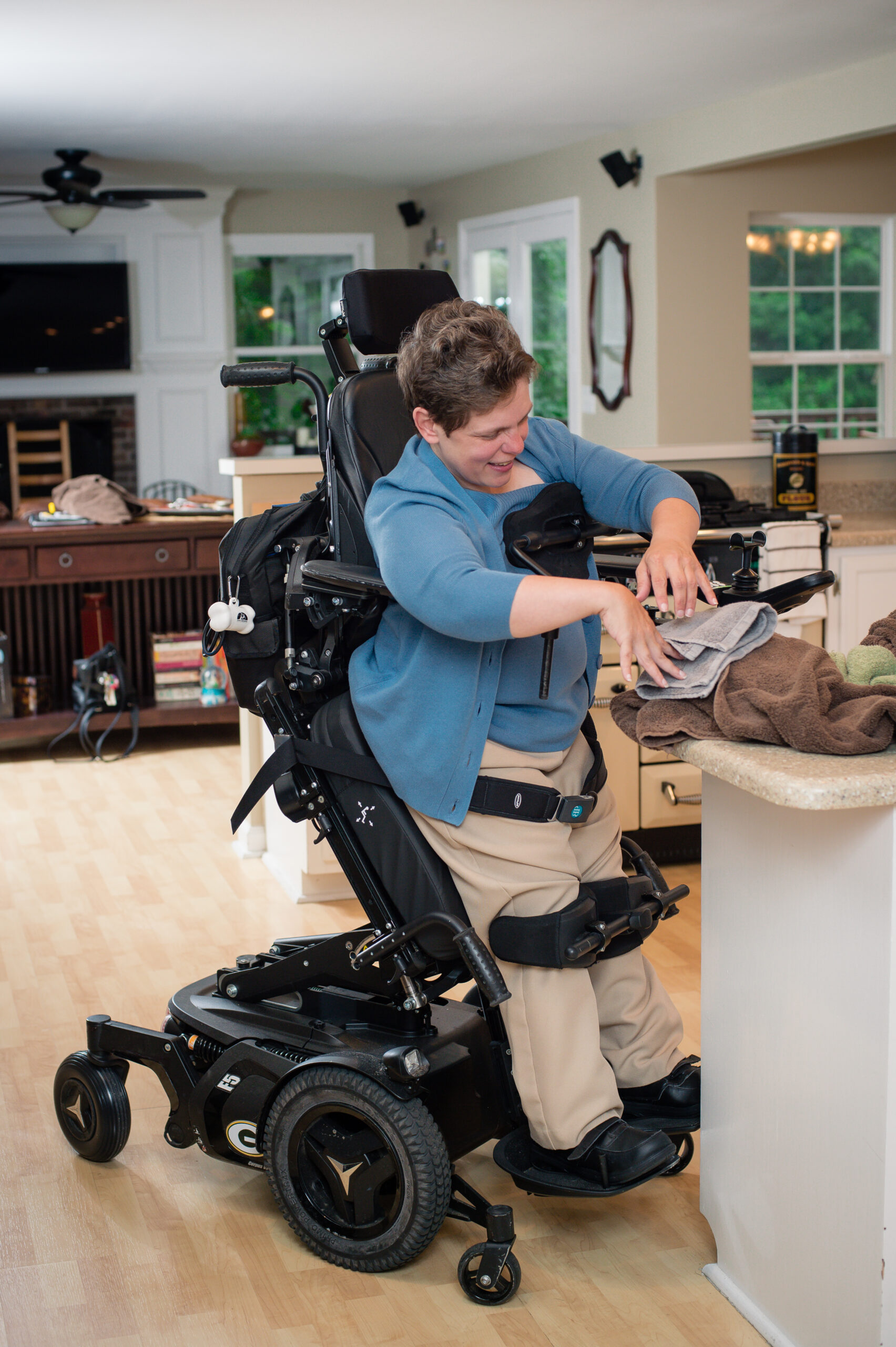 A smiling person using a power standing wheelchair folds towels at a kitchen counter in a bright, spacious room with large windows and wooden floors.