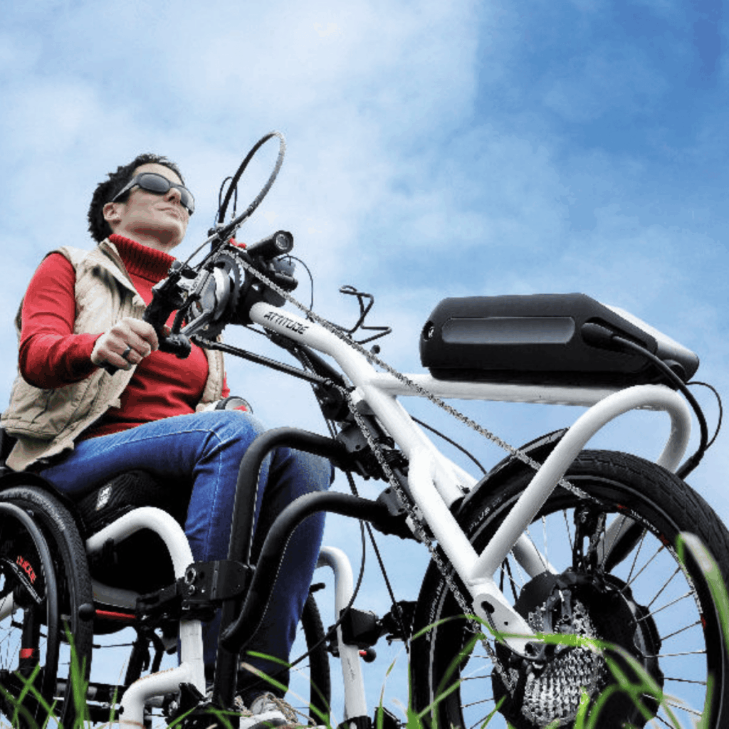 A woman in a manual wheelchair rides outdoors using a white handcycle-style power assist attachment. She wears sunglasses, a red turtleneck, and a tan vest, with a blue sky in the background.