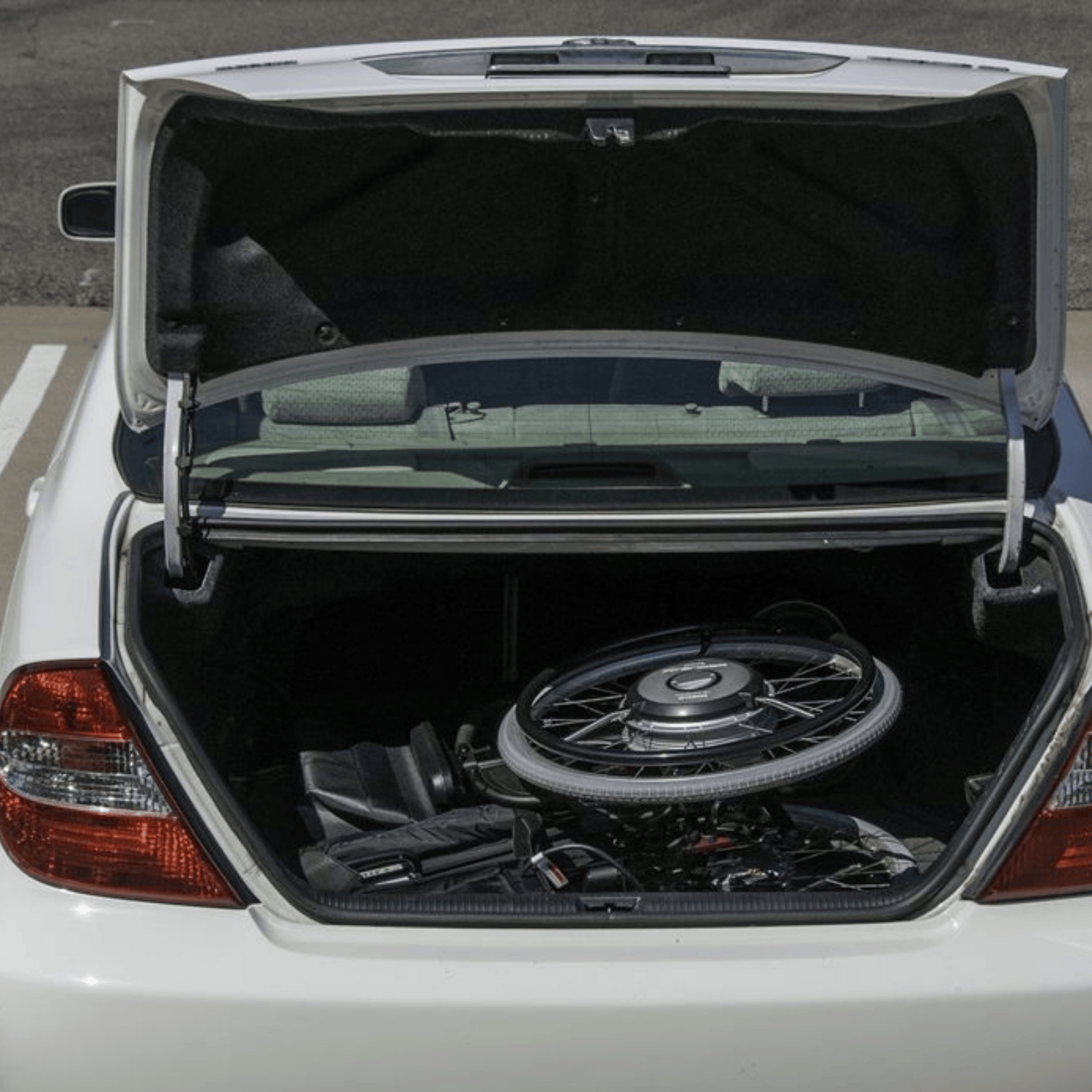 A lightweight manual wheelchair with power assist wheels is folded and stored neatly inside the trunk of a white sedan, showcasing its portability and compact design.