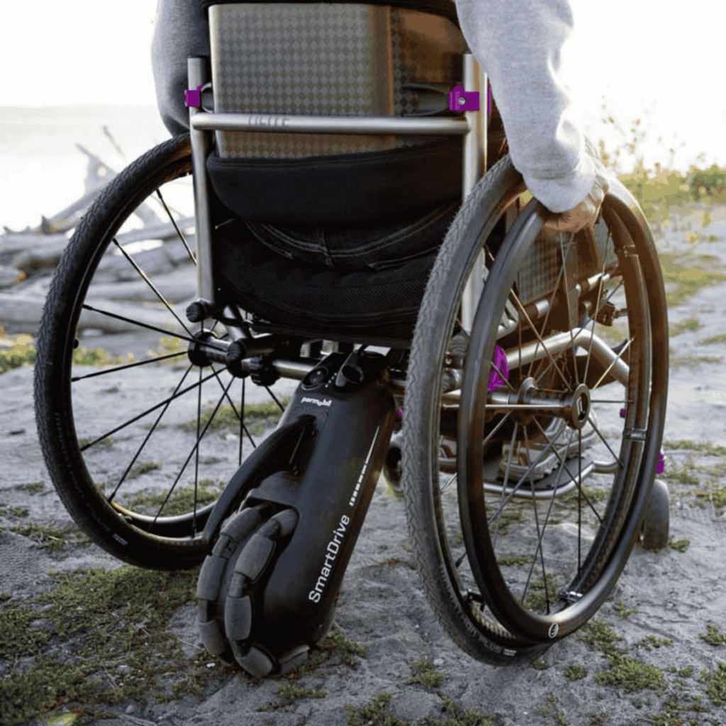 Close-up of the back of a manual wheelchair equipped with a SmartDrive power assist device on a sandy outdoor path. The user’s hand grips the wheel, with sunlight in the background.