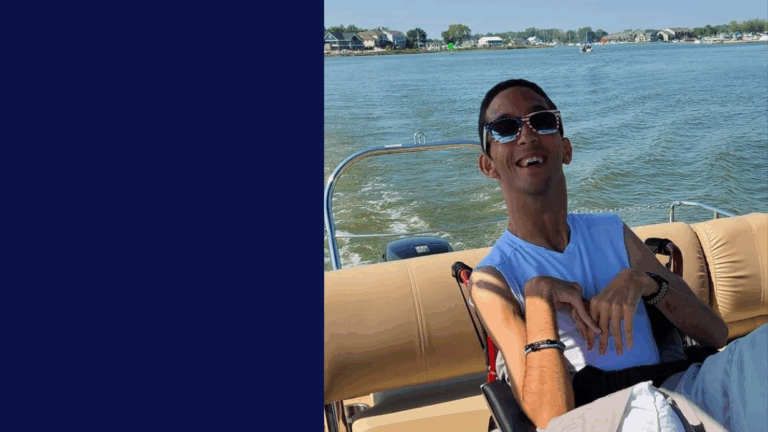 A man wearing sunglasses and a white sleeveless shirt smiles while sitting on a beige boat seat, with a lake and houses in the background on a sunny day, capturing Rickys Story.