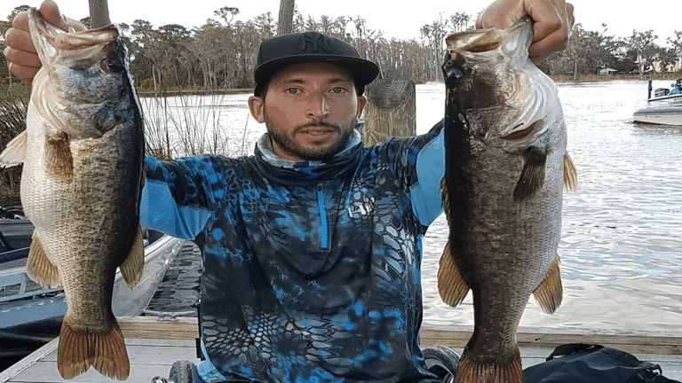 Sammys Story: A man in a blue patterned shirt and black cap holds up two large fish, one in each hand, on a dock by a lake with trees and a boat in the background.