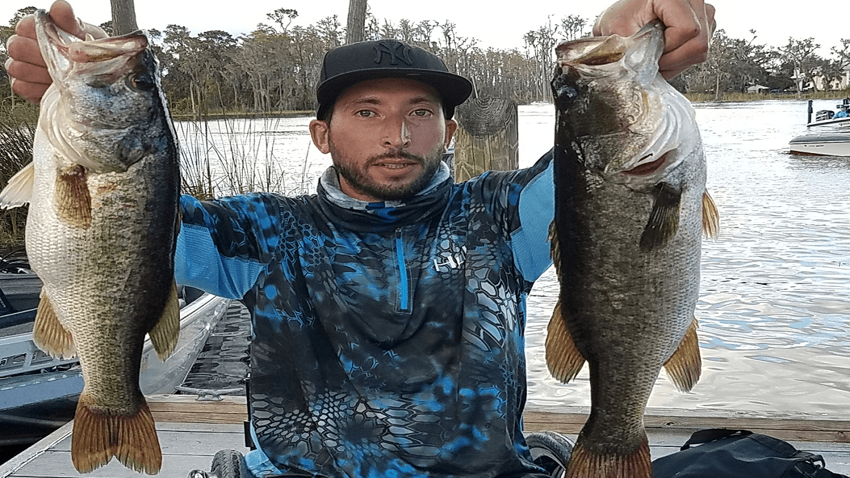 Sammys Story: A man in a blue patterned shirt and black cap holds up two large fish, one in each hand, on a dock by a lake with trees and a boat in the background.