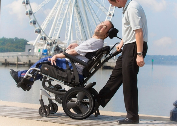 A man using adult mobility solutions smiles as he is guided by another man along a boardwalk near a body of water, with a large Ferris wheel in the background.