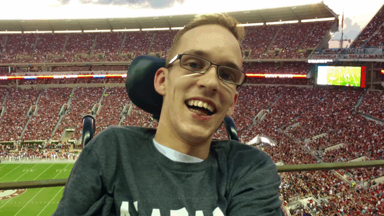 Seths Story: A young man wearing glasses and a gray shirt smiles at a crowded football stadium, with the stands filled with fans and the field visible in the background.