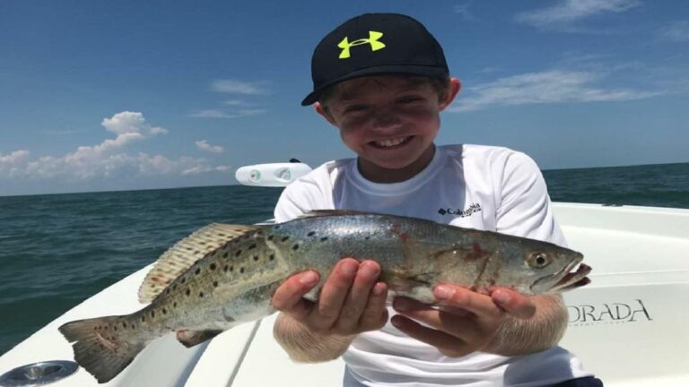 A smiling boy wearing a black cap and white shirt holds a large, spotted fish on a boat, with the ocean and blue sky in the background—capturing a moment from Stevens Story.