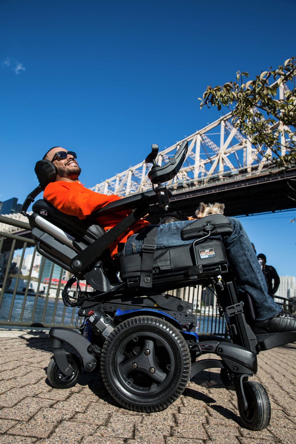A man in a power tilt wheelchair smiles while relaxing outdoors near a bridge on a sunny day, with blue sky and city scenery in the background.