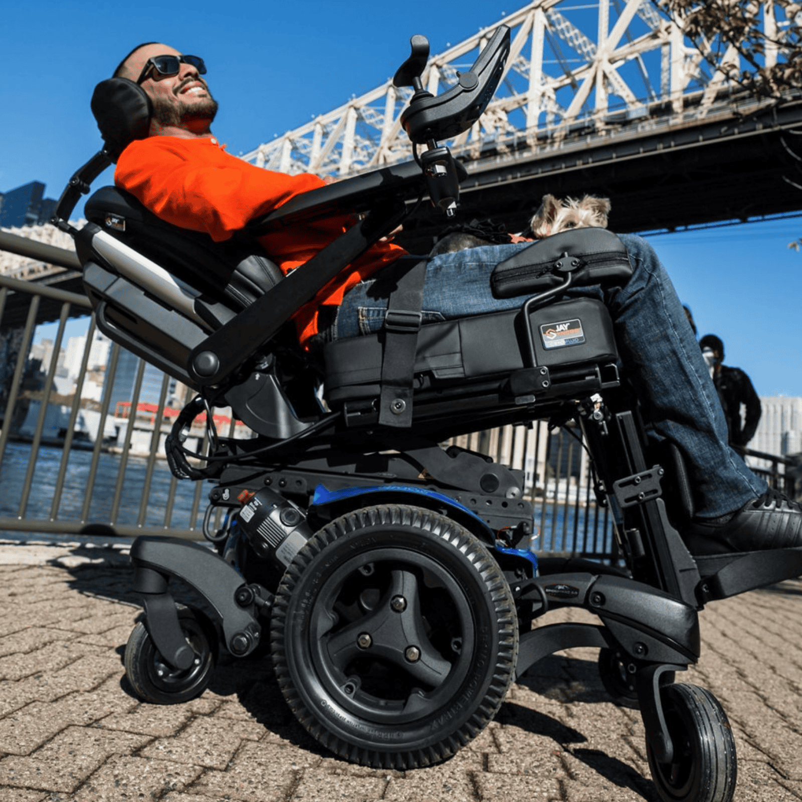 A smiling man in a bright red shirt relaxes in a fully tilted power wheelchair near a waterfront, with a large bridge in the background. The chair is reclined for comfort and support.