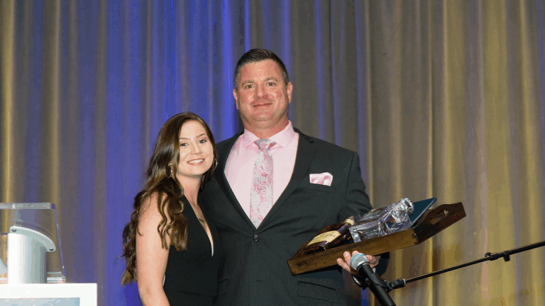 A woman in a black dress stands next to a man in a suit holding a gift tray, both smiling at an indoor event with a blue and gold curtain backdrop, capturing a moment from Todds Story.