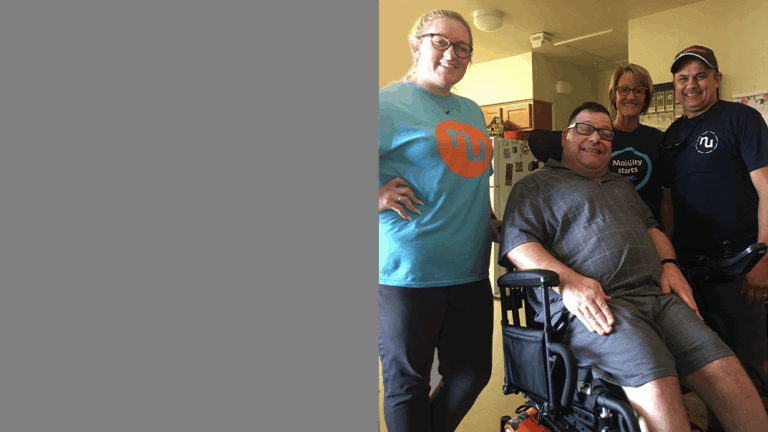 Four smiling adults pose together indoors; one man is seated in a motorized wheelchair, while the others stand around him. Everyone appears happy and relaxed in a home setting, sharing a moment from Todds Story.