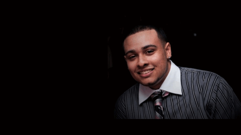 A man with short hair and trimmed facial hair smiles while wearing a striped dress shirt, a tie, and earrings. The background is dark, with most of the left side of the portrait in shadow.