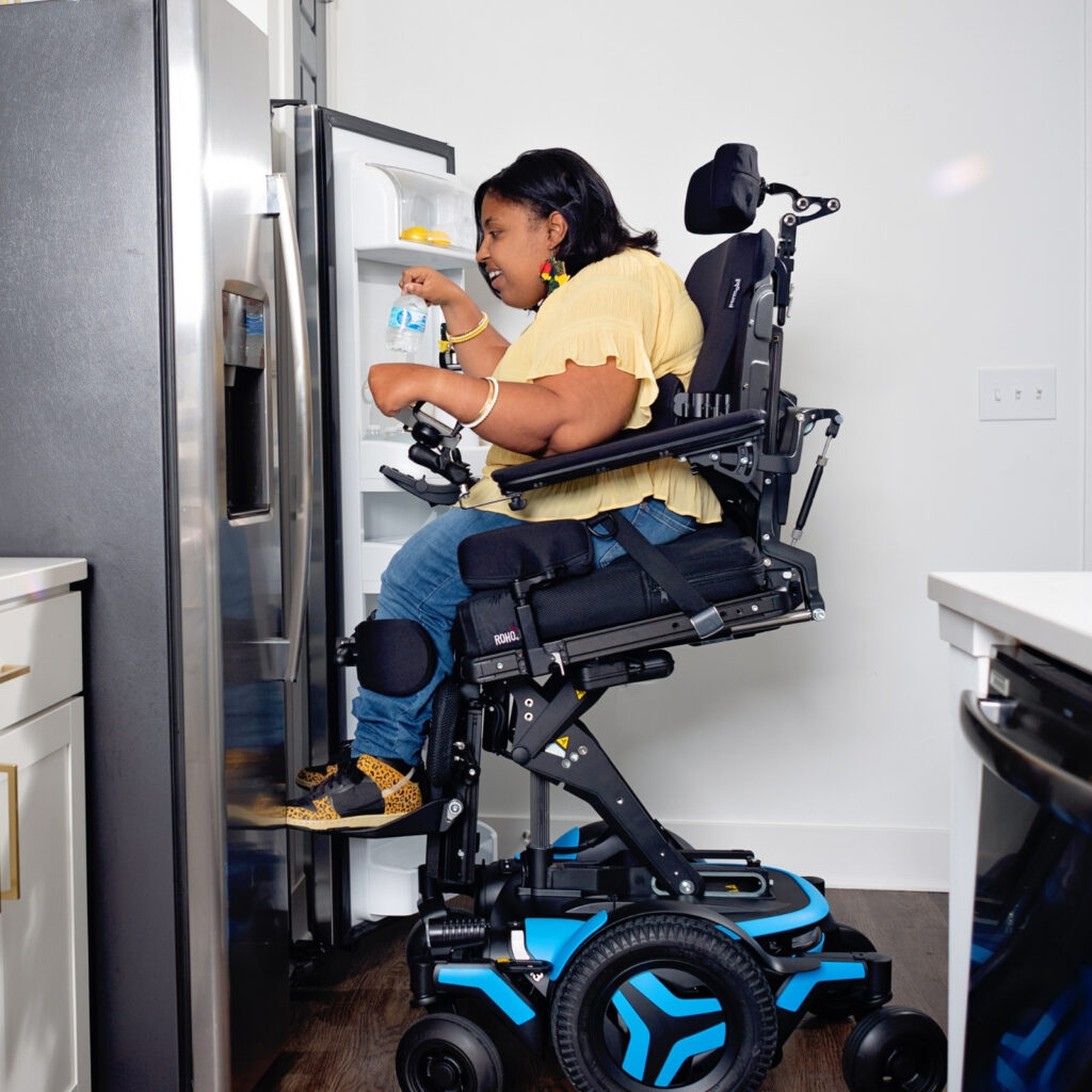 A woman using a motorized wheelchair with a Power Seat Elevator reaches for a water bottle in the refrigerator of a modern kitchen.