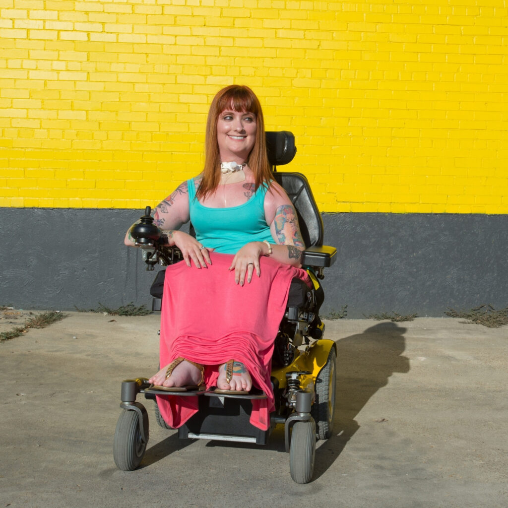 A woman with long brown hair, wearing a teal top and pink skirt, sits in a yellow power wheelchair—an example of adult mobility solutions—in front of a bright yellow brick wall with a dark gray base. She is smiling at the camera.