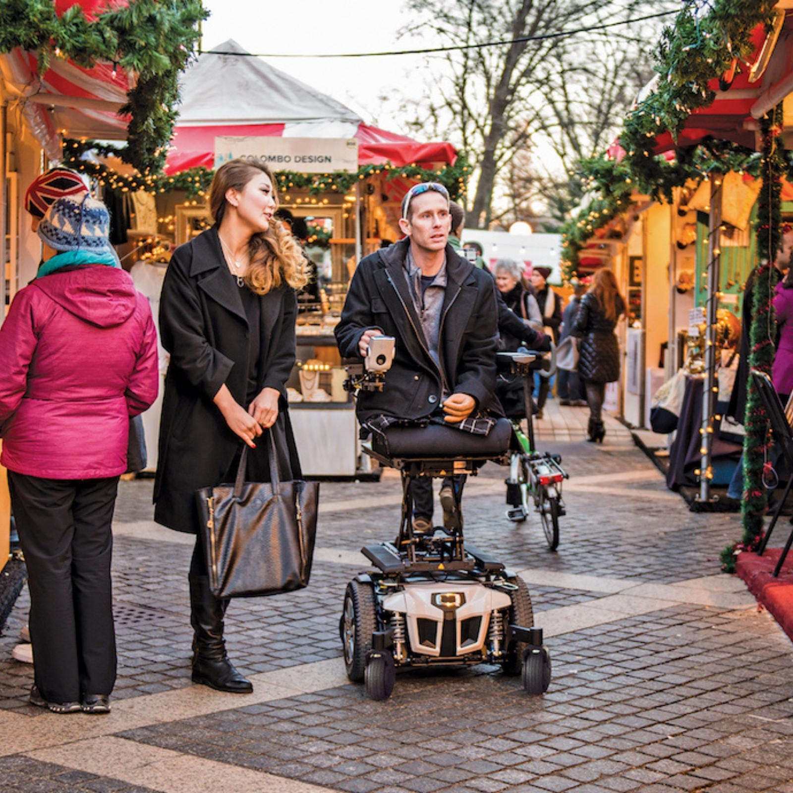A man using a Power Seat Elevator and a woman stand together at an outdoor holiday market. People walk nearby, and festive decorations adorn the stalls in the background.
