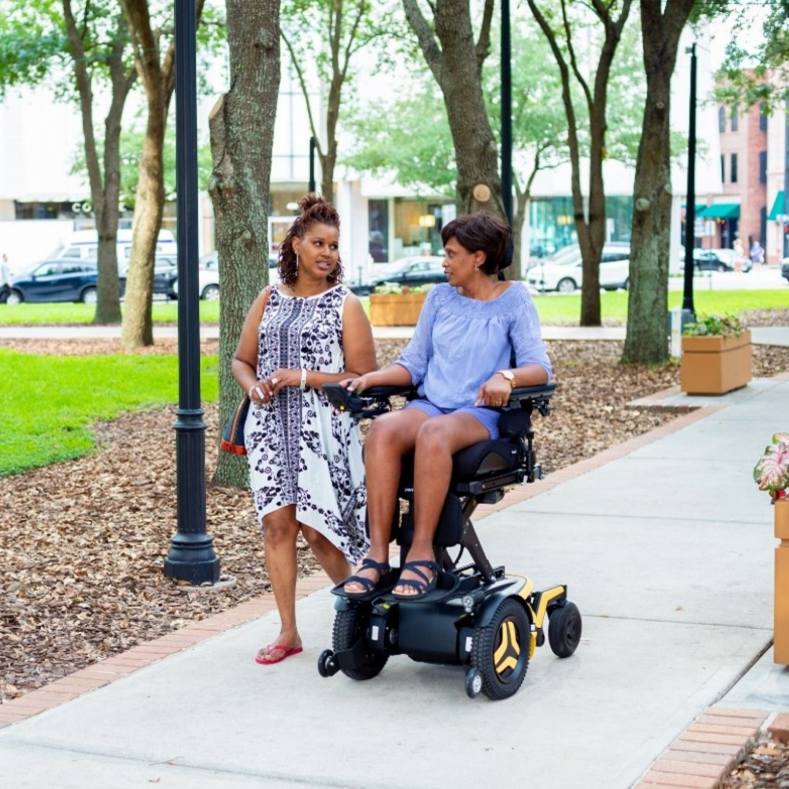 Two women enjoy a walk outdoors; one walks on the sidewalk while the other uses a power wheelchair with a Power Seat Elevator. They are smiling and engaged in conversation, surrounded by trees and parked cars.