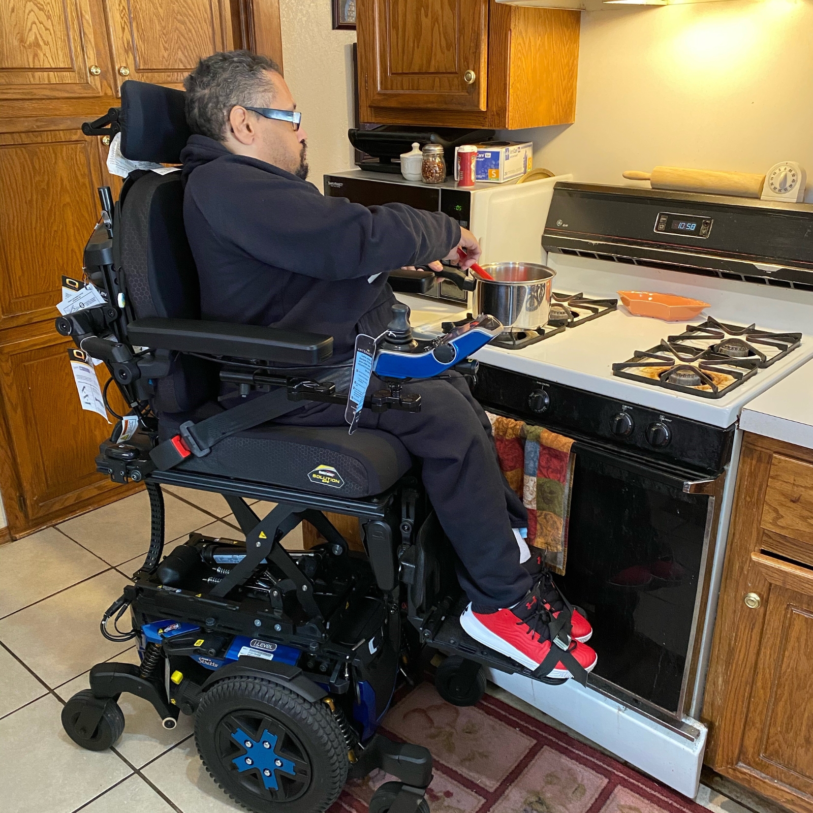 A man using a powered wheelchair with a Power Seat Elevator is cooking at a stove in the kitchen. He wears glasses, a dark tracksuit, and red-accented sneakers, stirring something in a pot over the burner.
