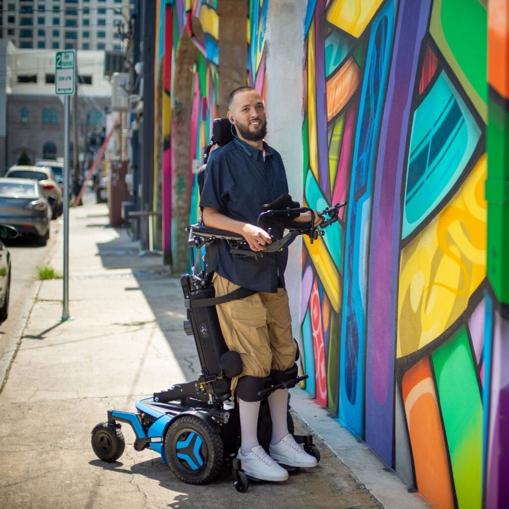 A man in a complex power standing wheelchair looks at a colorful mural on a city sidewalk, wearing a navy shirt, tan shorts, and white sneakers. Cars and buildings are visible in the background.