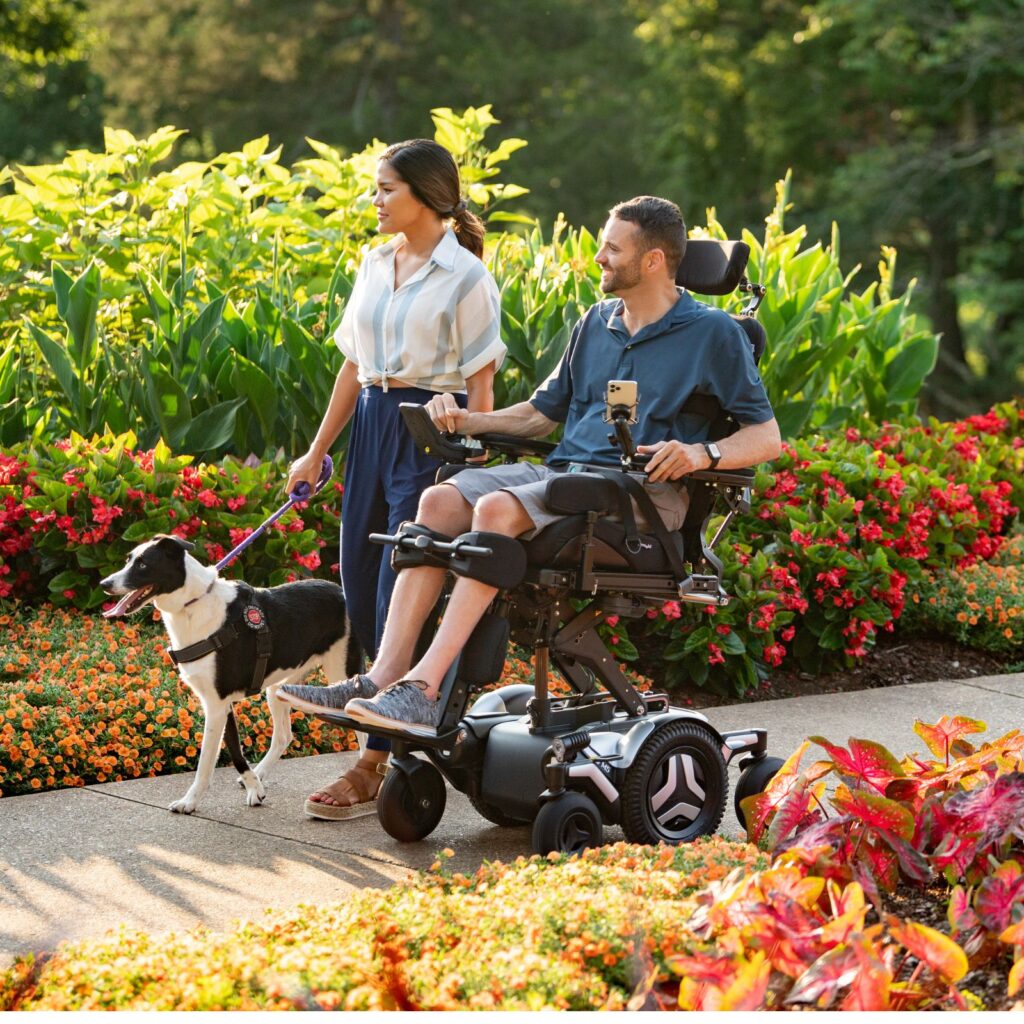 A woman walks beside a man in a motorized wheelchair with Complex Power Tilt on a garden path, holding the leash of a black and white dog. Colorful flowers and green plants surround them, and they appear happy and engaged.