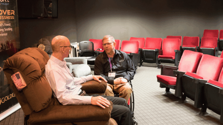 Two men are talking in a small theater with red seats. One man sits in a brown recliner while the other, featured in Jeffs Story, sits across from him. The theater seats behind them are empty.