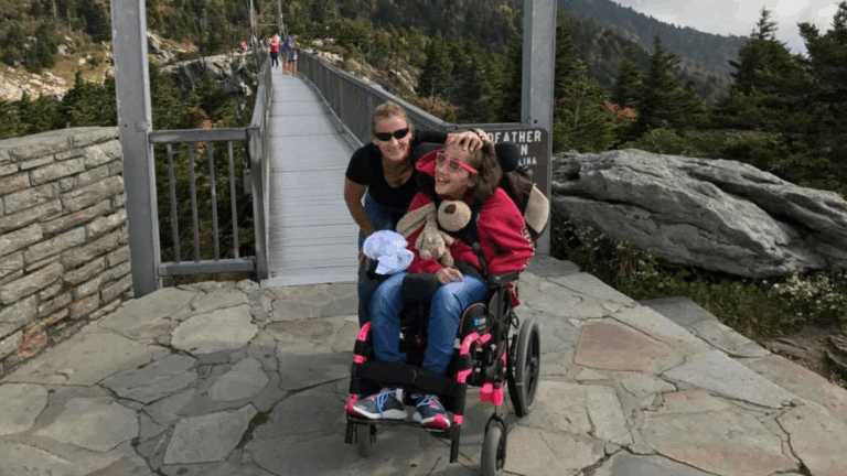 Lexis Story: A woman stands behind a smiling girl in a wheelchair at the entrance to a suspension bridge, surrounded by trees and mountains under a cloudy sky.