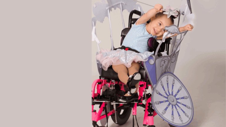 Avery Joy, dressed as a princess, smiles and poses with her arms raised. Her wheelchair, decorated like a fairy-tale carriage with pink and purple accents, tells an inspiring personal story of joy and imagination.