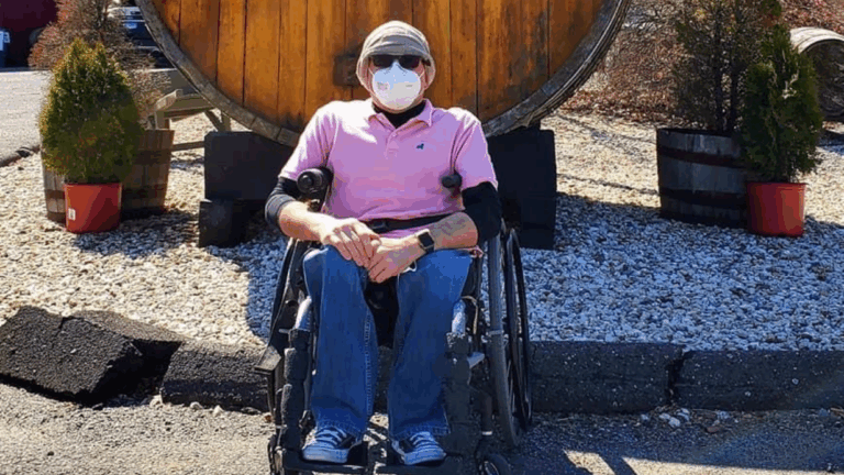 Steves Story: A person wearing a mask, sunglasses, hat, and pink shirt sits in a wheelchair outdoors in front of a large wooden barrel, with small potted trees on either side.
