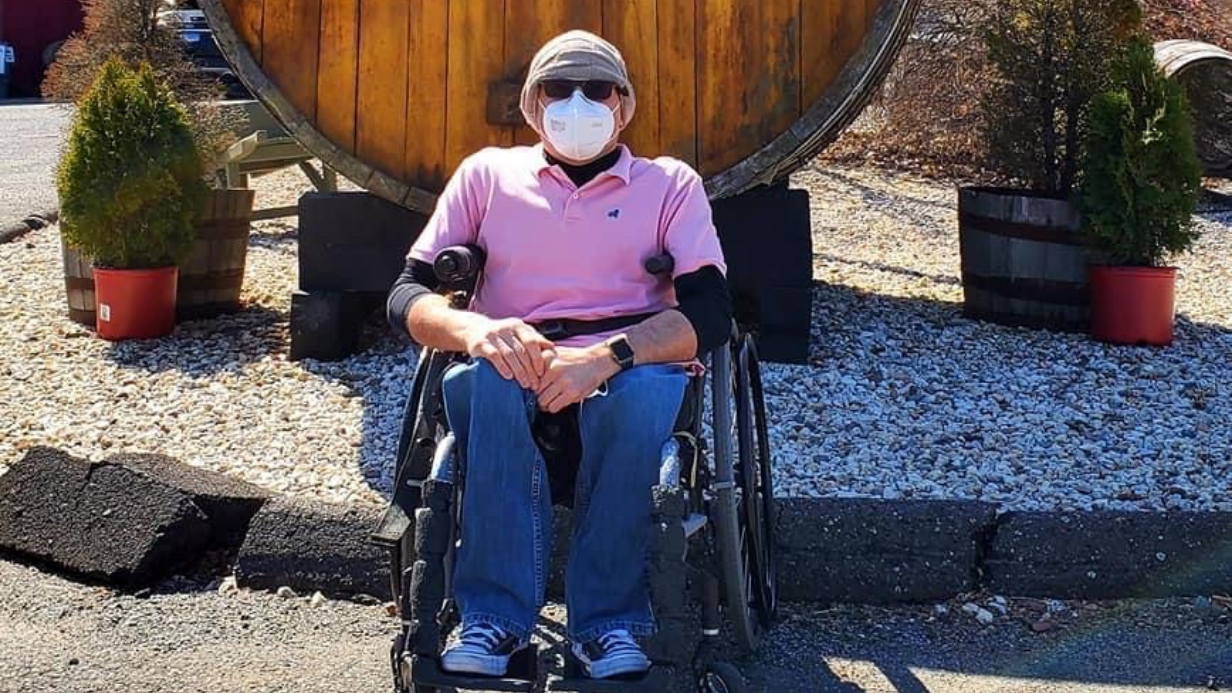 Steves Story: A person wearing a mask, sunglasses, hat, and pink shirt sits in a wheelchair outdoors in front of a large wooden barrel, with small potted trees on either side.