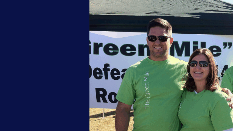 Two people wearing matching light green The Green Mile shirts stand smiling in front of a large white sign outdoors on a sunny day, sharing Jills Story. The left side of the image is covered by a dark blue rectangle.