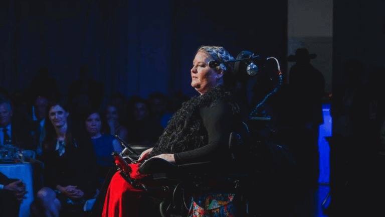 A woman using a wheelchair speaks onstage at an event, illuminated by a spotlight, as an audience listens attentively in a dimly lit room.