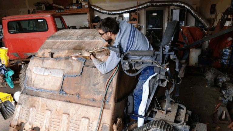 Chriss Story: A person wearing safety glasses uses a welding torch on a large, rusty metal bucket attachment in a garage workshop, sitting on a tracked chair. Tools and equipment are scattered around the space.