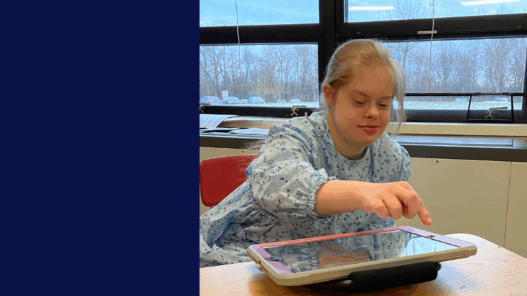 Hannahs Story: A girl with Down syndrome sits at a desk in a classroom, using a tablet with her finger. She is wearing a blue patterned top and is focused on the screen, with large windows and trees in the background.