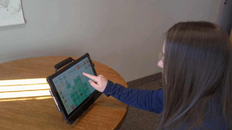 Laurens Story: A woman with long brown hair sits at a round wooden table, using a tablet with a colorful communication app. She touches the screen with her finger.