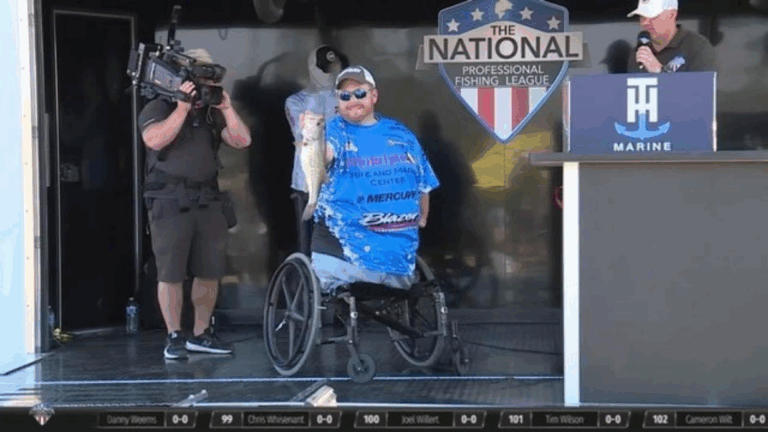 A smiling angler in a wheelchair proudly holds a large fish on stage at a National Professional Fishing League event, sharing Hunters Story while being filmed and interviewed by event staff.