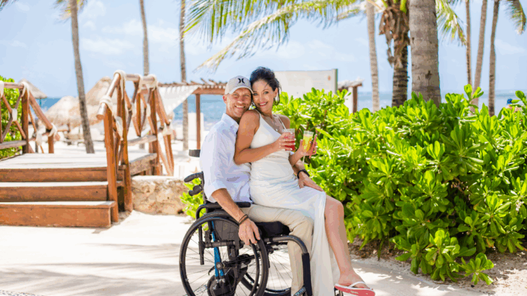 A smiling couple enjoys a sunny beach day; the woman sits on a mans lap as he relaxes in his wheelchair, both holding drinks. Palm trees, greenery, and a sandy path with ocean in the background capture Kriss Story.