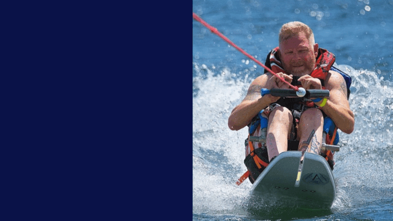 A man with a focused expression is seated on a waterski board, gripping a tow rope as he skims across the water in Davids Story, creating splashes. The left side of the image is solid dark blue.
