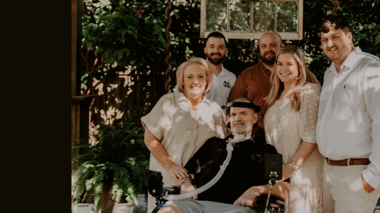 Six adults, including a man with medical equipment, are posing together outdoors in a sunlit garden. Some are standing while one is seated, and everyone is smiling as they share a moment from Kevins Story.