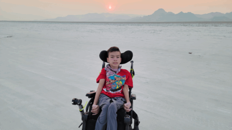 A boy in a wheelchair sits on a vast, flat sandy landscape at sunset, with a mountain range in the distance and a soft orange sky overhead, capturing a moment from Xaviers Story.