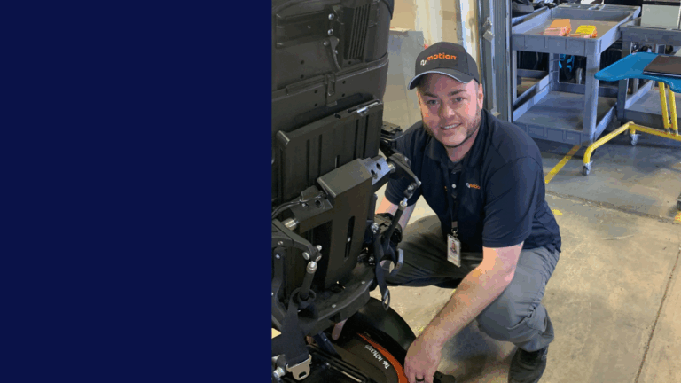 Rodneys Story: A man in a black shirt and cap labeled eMotion kneels while working on a power wheelchair in an industrial setting, smiling at the camera.