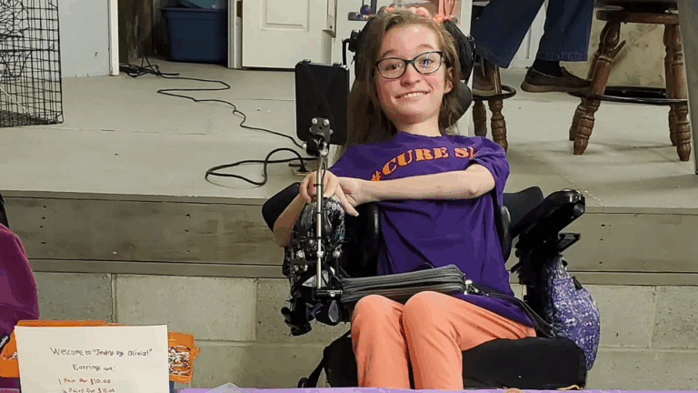A smiling young woman with glasses sits in a wheelchair behind a table, wearing a purple Cure SMA T-shirt and peach pants. A handwritten sign and some items are on the table, highlighting Olivias Story.