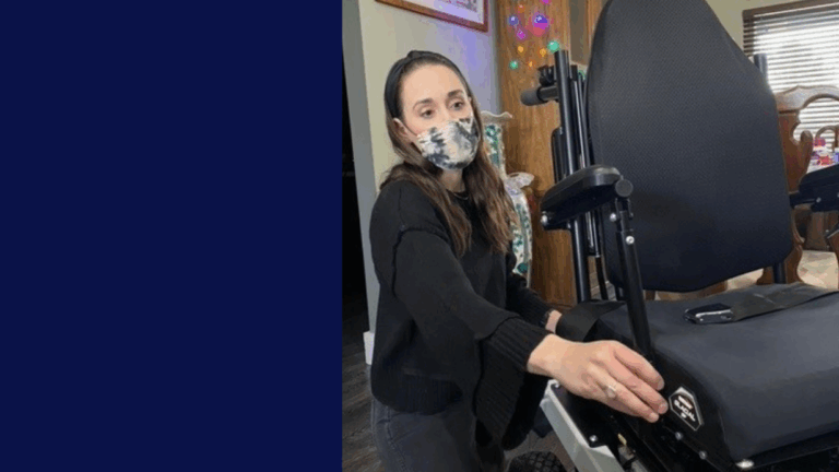 A woman wearing a patterned face mask and black sweater adjusts a black wheelchair indoors in Caitlins Story. Colorful lights and a window with blinds illuminate the background.