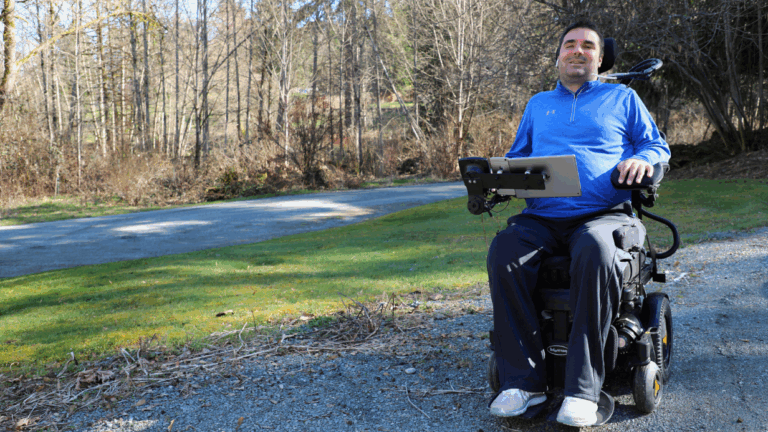 Tylers Story: A man in a blue jacket and dark pants sits in a power wheelchair with a mounted tablet, smiling outdoors on a sunny day with trees and grass in the background.