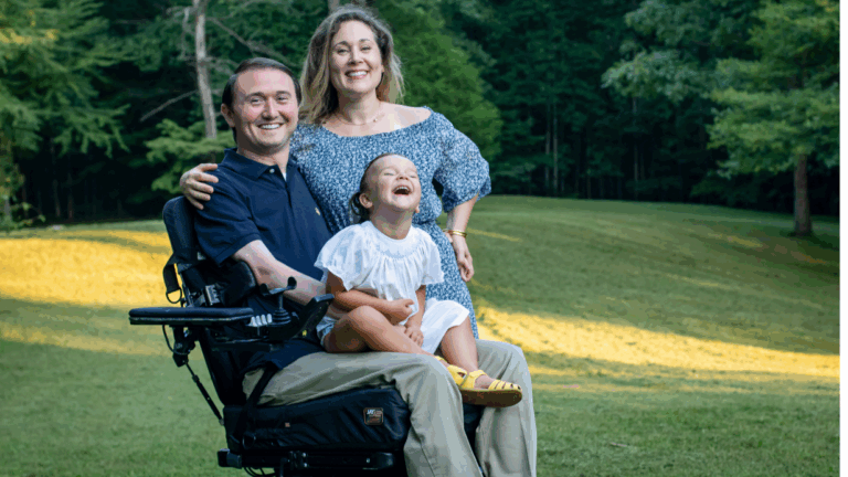 A smiling man in a wheelchair sits on grass with a laughing young girl on his lap and a smiling woman standing behind them, all outdoors in a green park—capturing a joyful moment from Joshs Story.