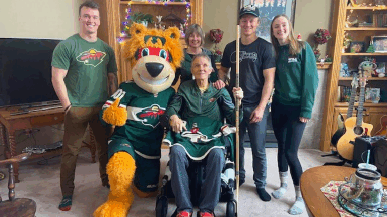 A group of five smiling people, one using a wheelchair, pose indoors with a person in a fuzzy orange hockey mascot costume. Everyone is wearing Minnesota Wild apparel in shades of green, surrounded by homey decorations.